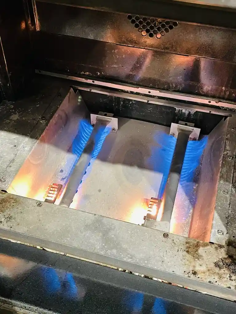A close-up photo shows the interior of a stainless steel oven with two burners ignited, emitting bright blue flames and…