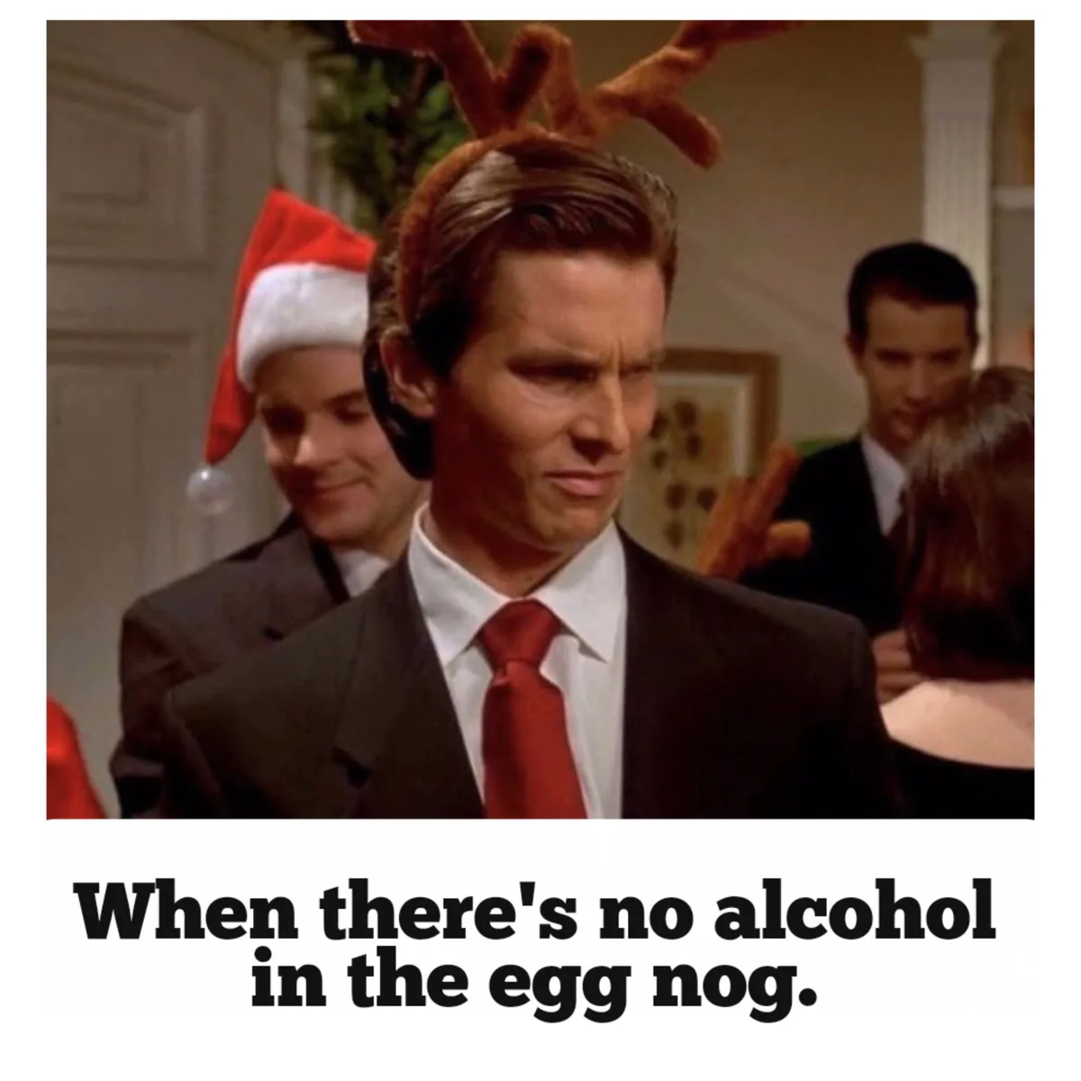 A man in a suit and red tie wears brown reindeer antlers and has a disgusted, skeptical expression. Behind him, another man…