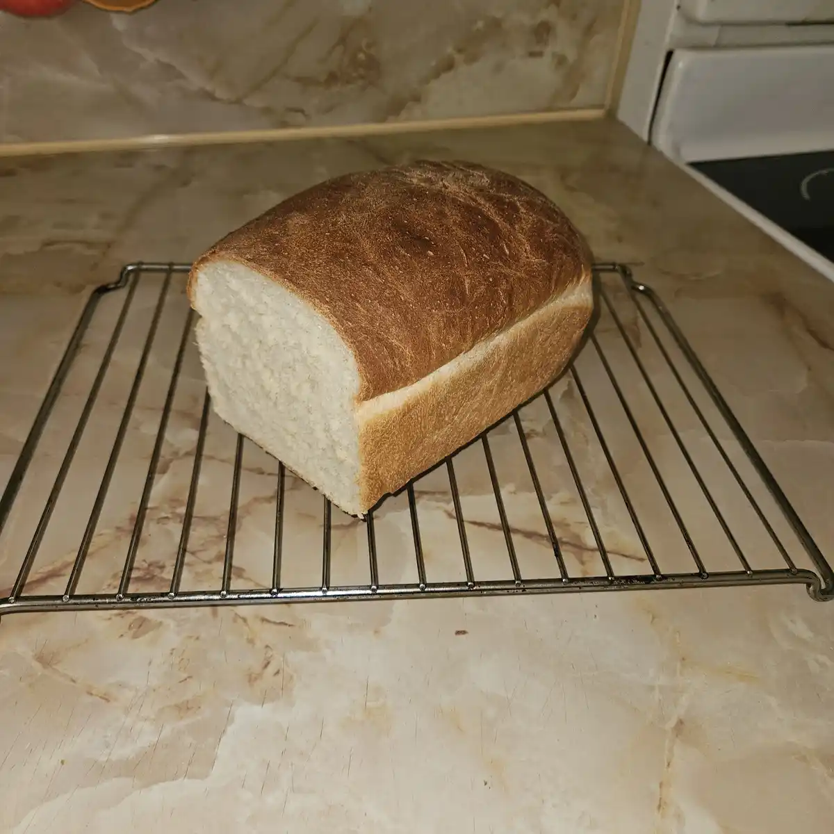 A freshly baked loaf of bread with a golden-brown crust sits on a metal cooling rack on a beige marble countertop. The bread…