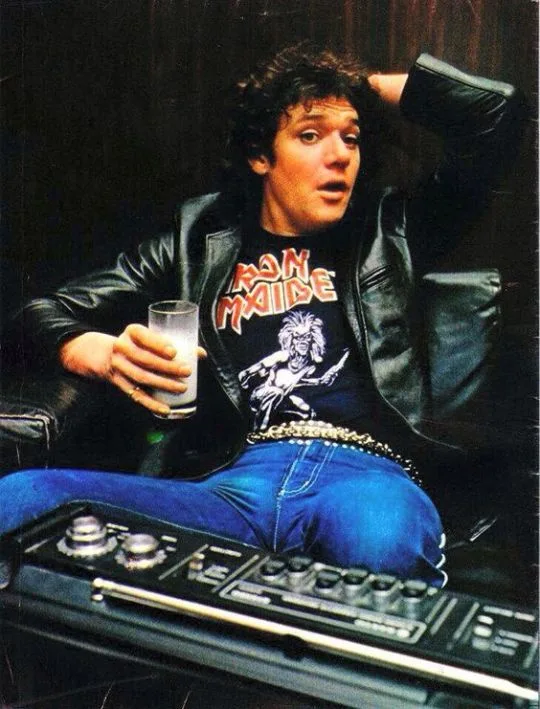 A man with curly hair wearing a black leather jacket and blue jeans sits on a vintage stereo console, holding a glass of…