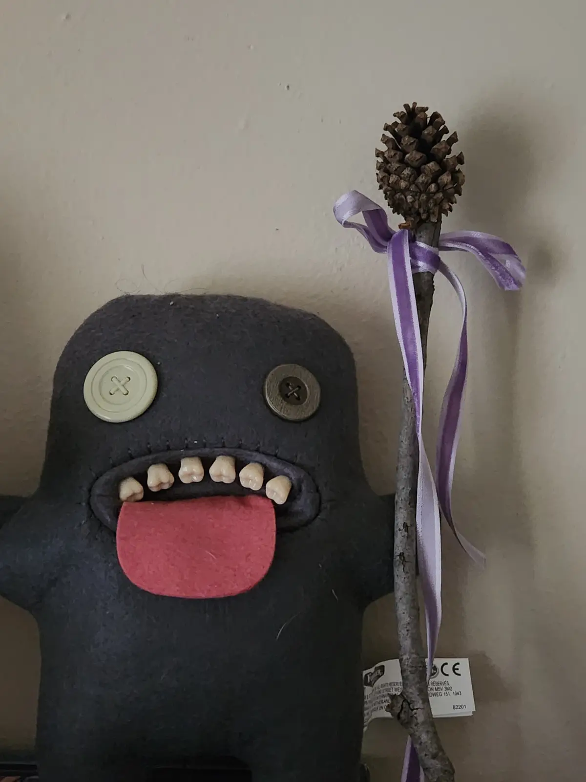 A handmade dark grey felt monster with button eyes, a row of small teeth, and a red tongue sticking out, holding a stick…