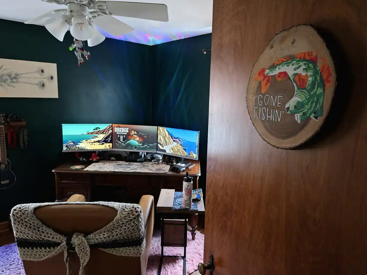 A view into a gaming room with three monitors displaying the game 'Dredge' and a cozy chair draped in a crochet blanket. On…