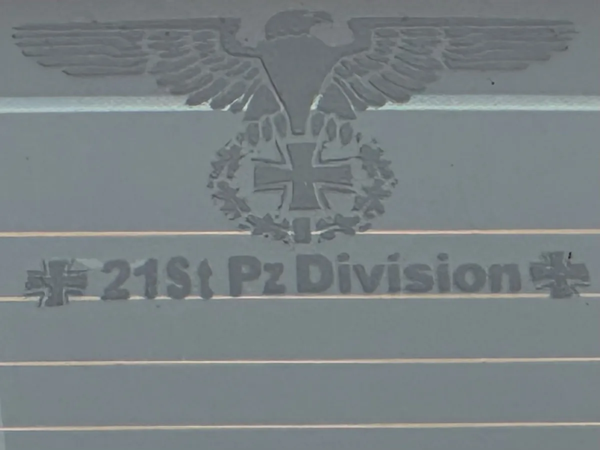 A close-up of a gray metal plaque featuring a raised emblem of a Nazi eagle clutching a swastika wreathed Iron Cross, with…