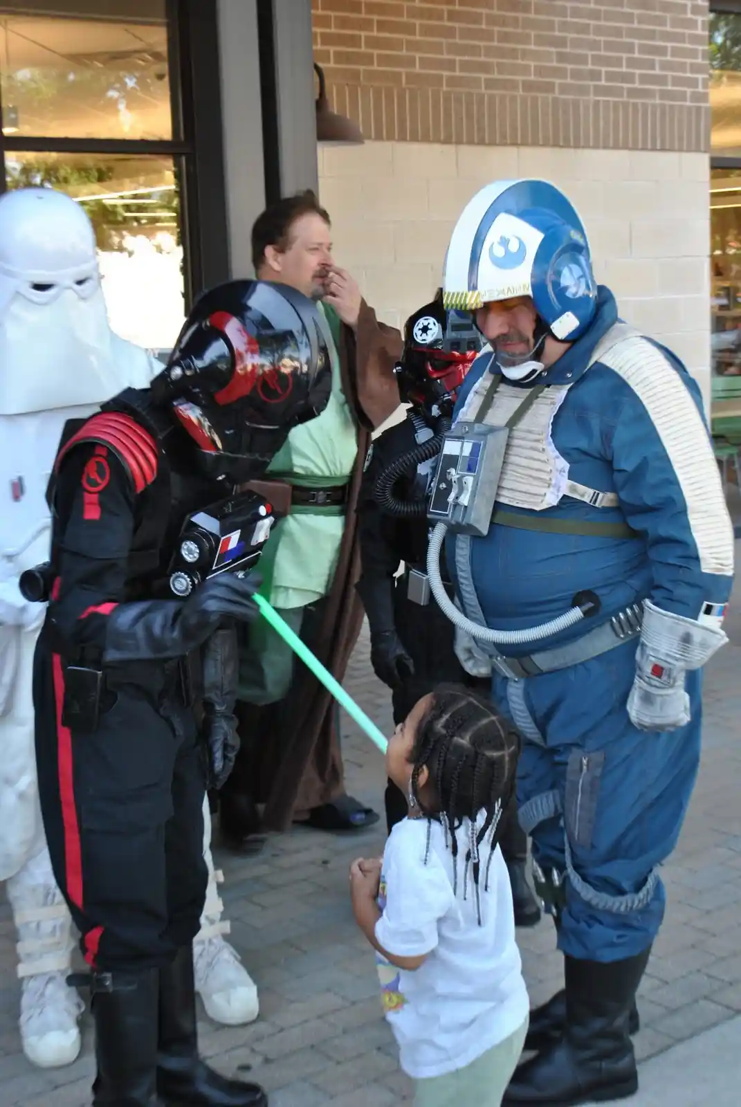 A young child in a white shirt looks up at a person in a blue and white Star Wars pilot costume holding a green lightsaber…