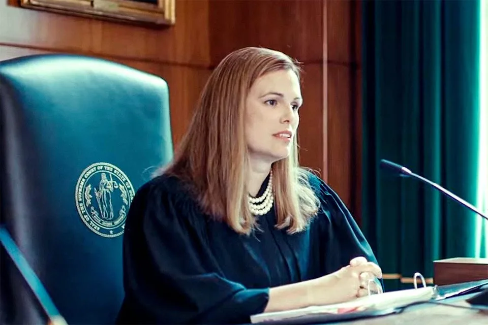 A woman in a black judicial robe sits in a high-backed blue chair with a court seal on it, looking slightly to her right…
