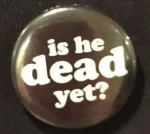 A close-up of a round, glossy button with white text on a dark background that reads 'is he dead yet?'. The button appears…
