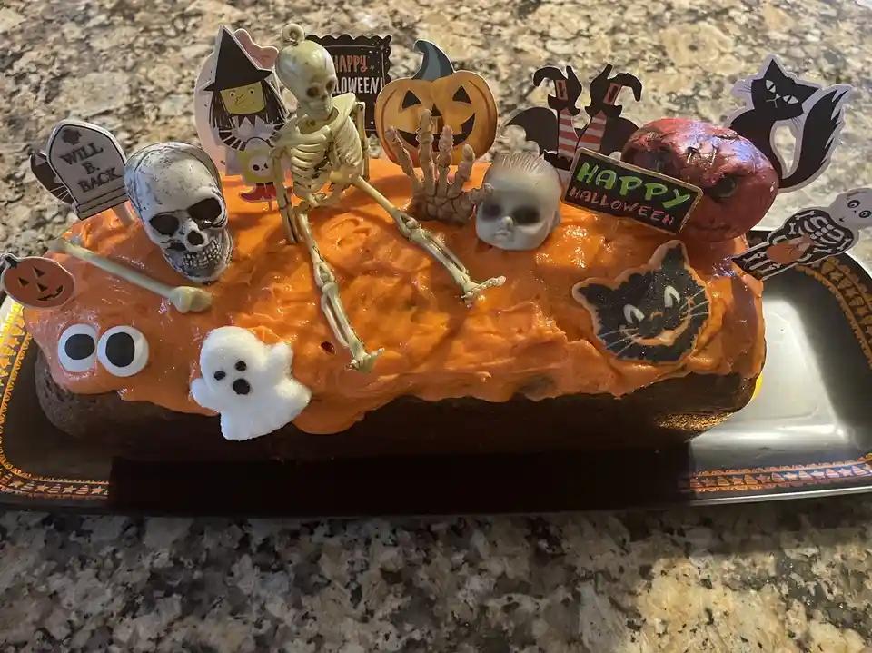 An orange-frosted Halloween cake is heavily decorated with spooky toppers including a skeleton, skulls, a baby doll head…