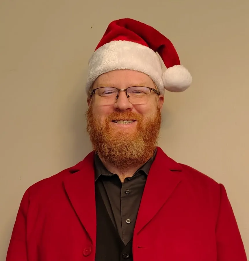 A man with a red beard and glasses wears a red coat, a black shirt, and a red-and-white Santa hat, smiling at the camera…