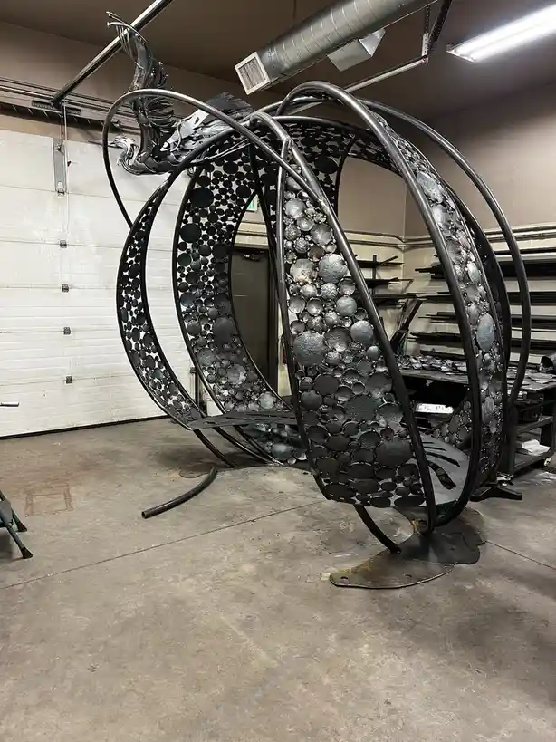 A large, intricate metal sculpture resembling a phoenix with curved wings and a body made of circular metal discs is…