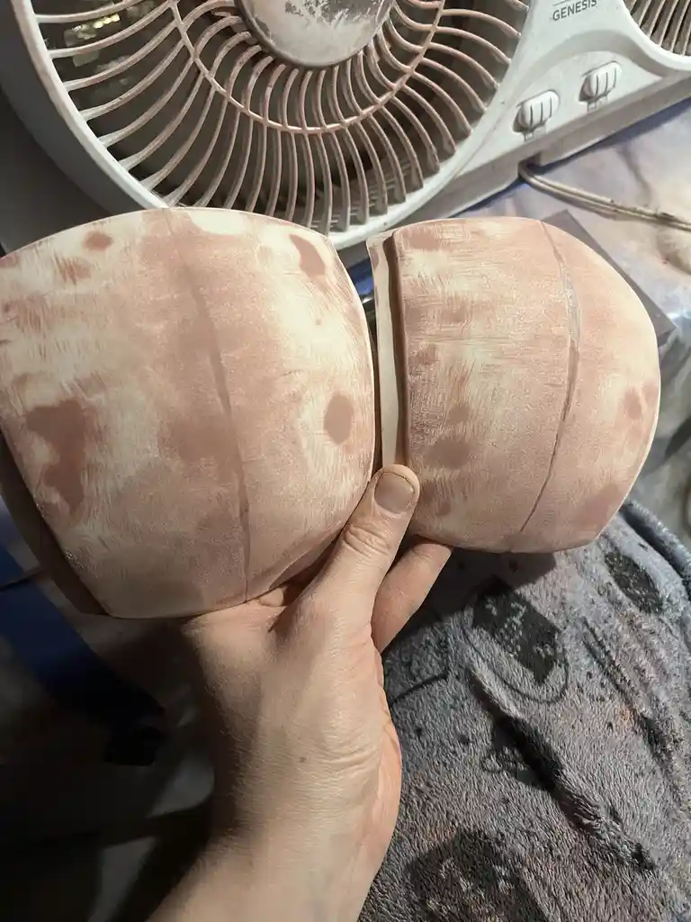 A hand holds two half-spheres painted to resemble the moon's surface, showing craters and texture, with a white box fan…
