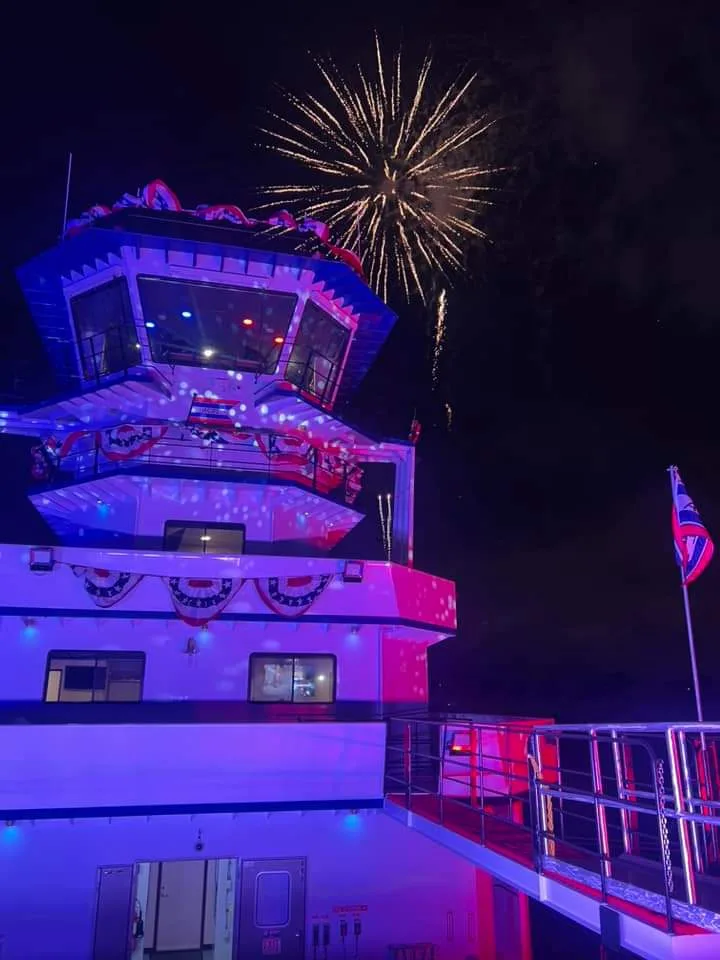 A nighttime photo of a multi-level boat decorated with patriotic bunting and illuminated by purple and pink lights, with…