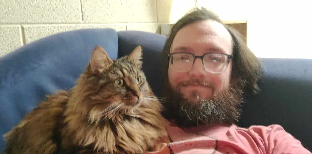 A man with glasses and a beard sits on a blue couch next to a large, fluffy brown tabby cat. The cat looks away from the…