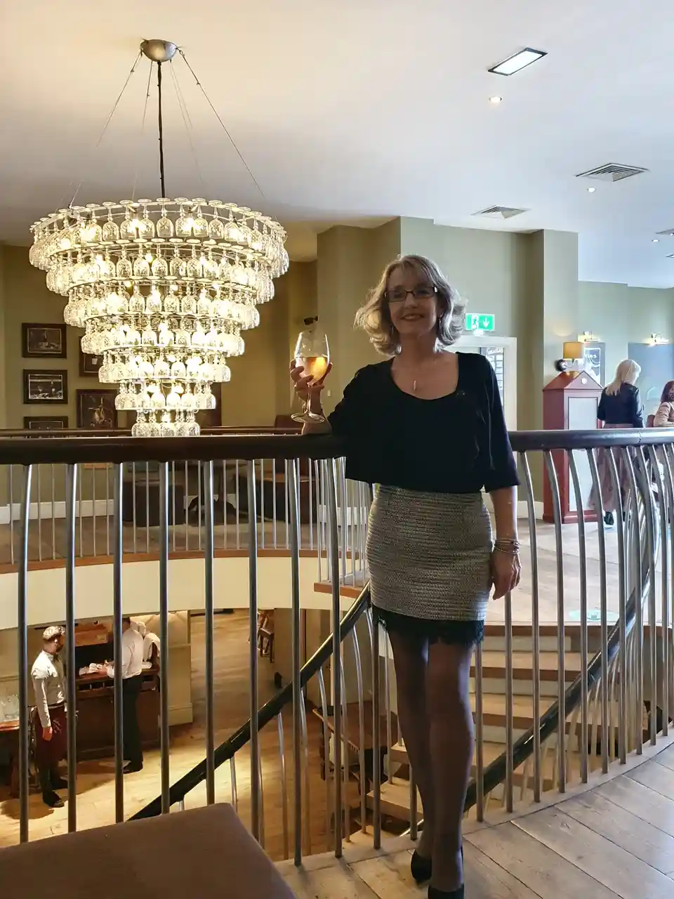A woman with short blonde hair and glasses stands smiling on a wooden staircase landing, holding a glass of white wine. She…