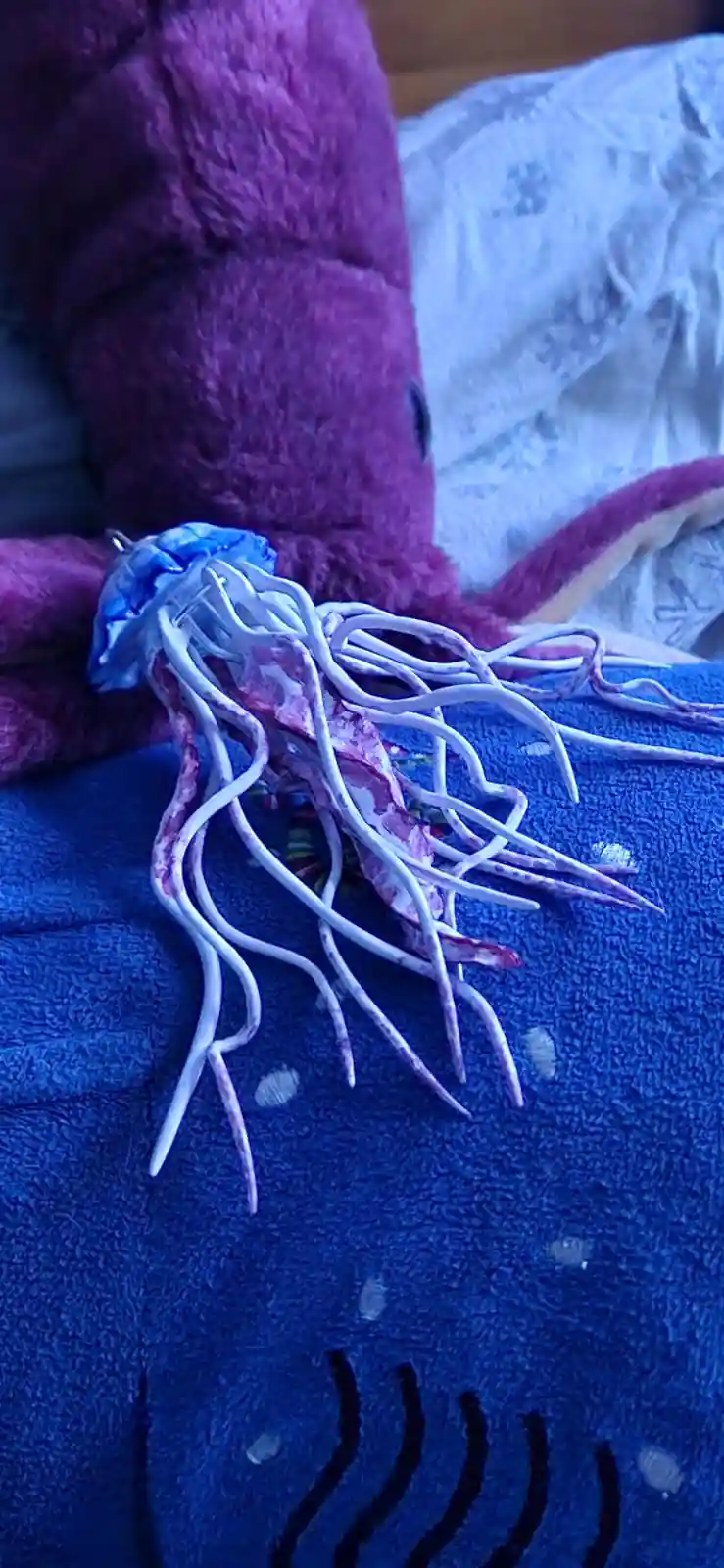 A plush toy resembling a jellyfish with a blue dome and long, white, tentacle-like appendages rests on a blue blanket with…