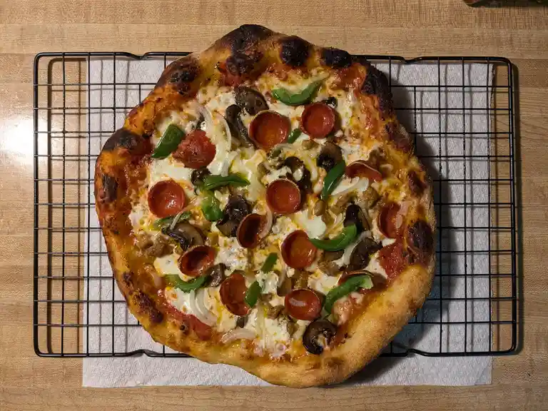 A freshly baked pizza with pepperoni, green peppers, mushrooms, onions, and sausage sits on a black wire cooling rack over a…