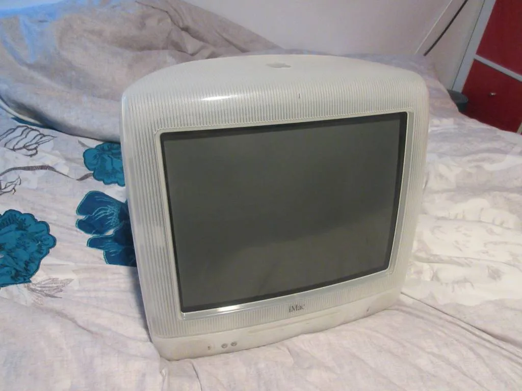 A vintage white iMac G3 computer with a ribbed plastic casing sits upright on a bed with floral-patterned sheets. The screen…