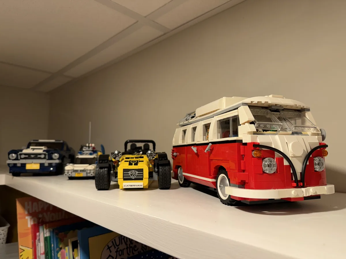 A white shelf holds four detailed LEGO car models: a blue classic car, a white police car, a yellow Caterham race car, and a…