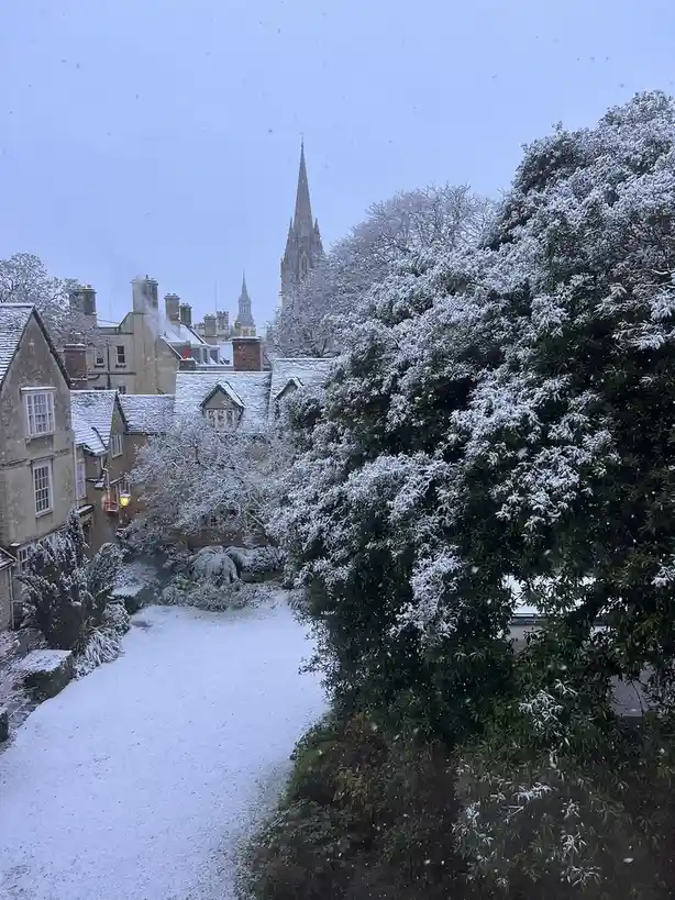 A snowy scene in a quaint town with stone buildings, snow-covered rooftops, and trees, with a church spire visible in the…