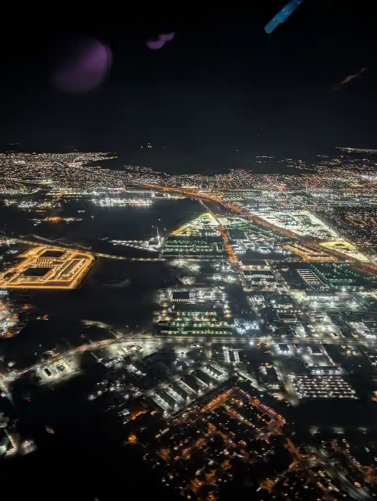 An aerial night view from an airplane window shows a sprawling, brightly lit airport complex with runways, terminals, and…