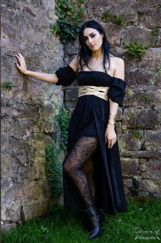 A woman with long dark hair stands against a mossy stone wall, wearing a black off-the-shoulder dress with a gold belt…