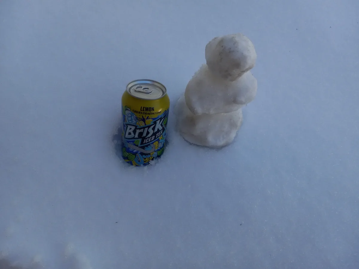 A yellow can of Brisk Iced Tea Lemon sits partially buried in snow next to a small, three-sphere snowman, both on a snowy…