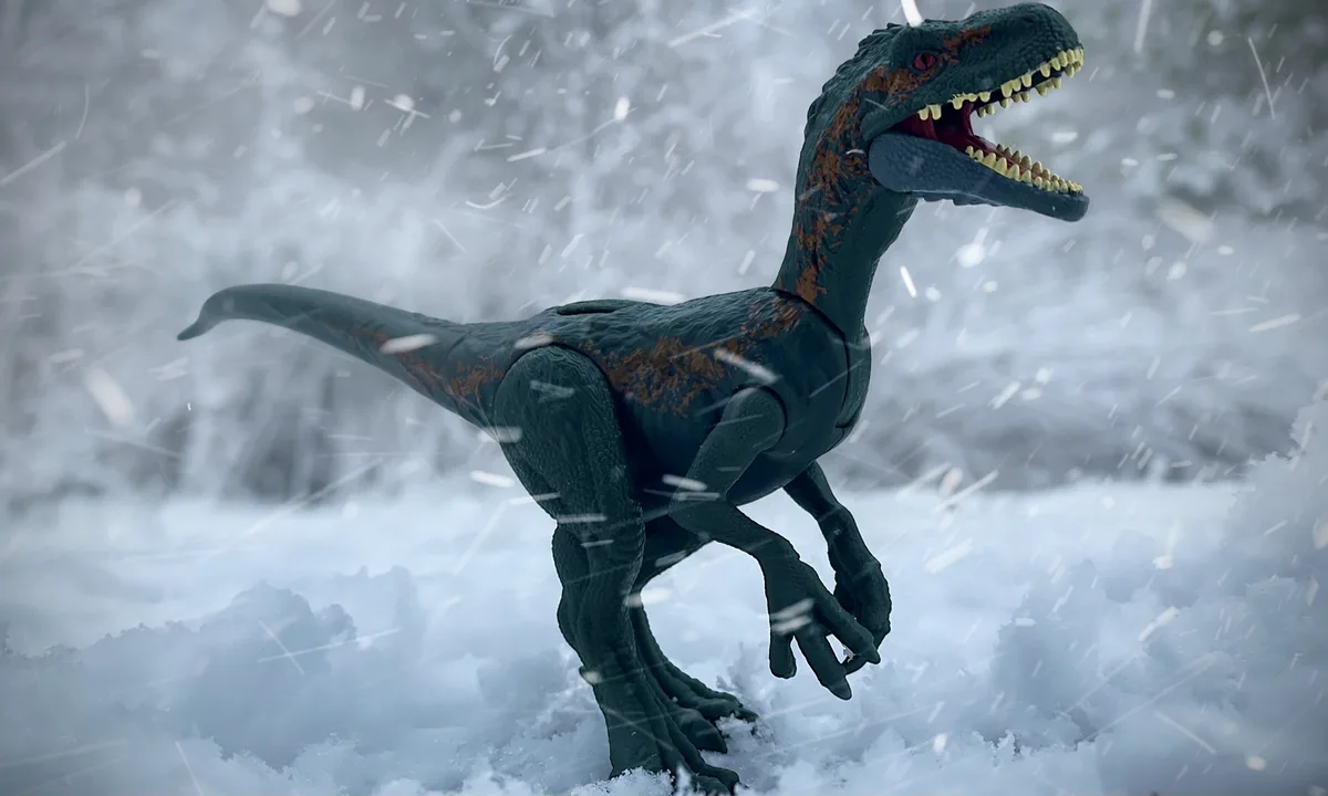 A dark green toy dinosaur with orange markings stands in deep snow as snowflakes fall around it, its mouth open in a roar…