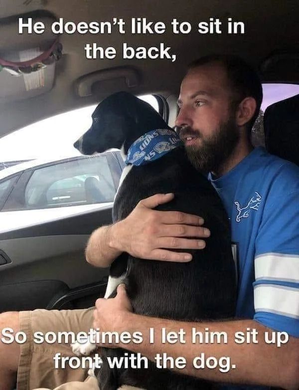 A man with a beard wearing a blue Detroit Lions t-shirt is sitting in the front passenger seat of a car, holding a black and…
