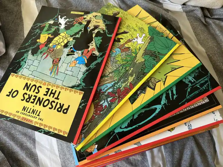 A fanned-out stack of Tintin comic books, with 'Prisoners of the Sun' on top, showing colorful covers with characters in…