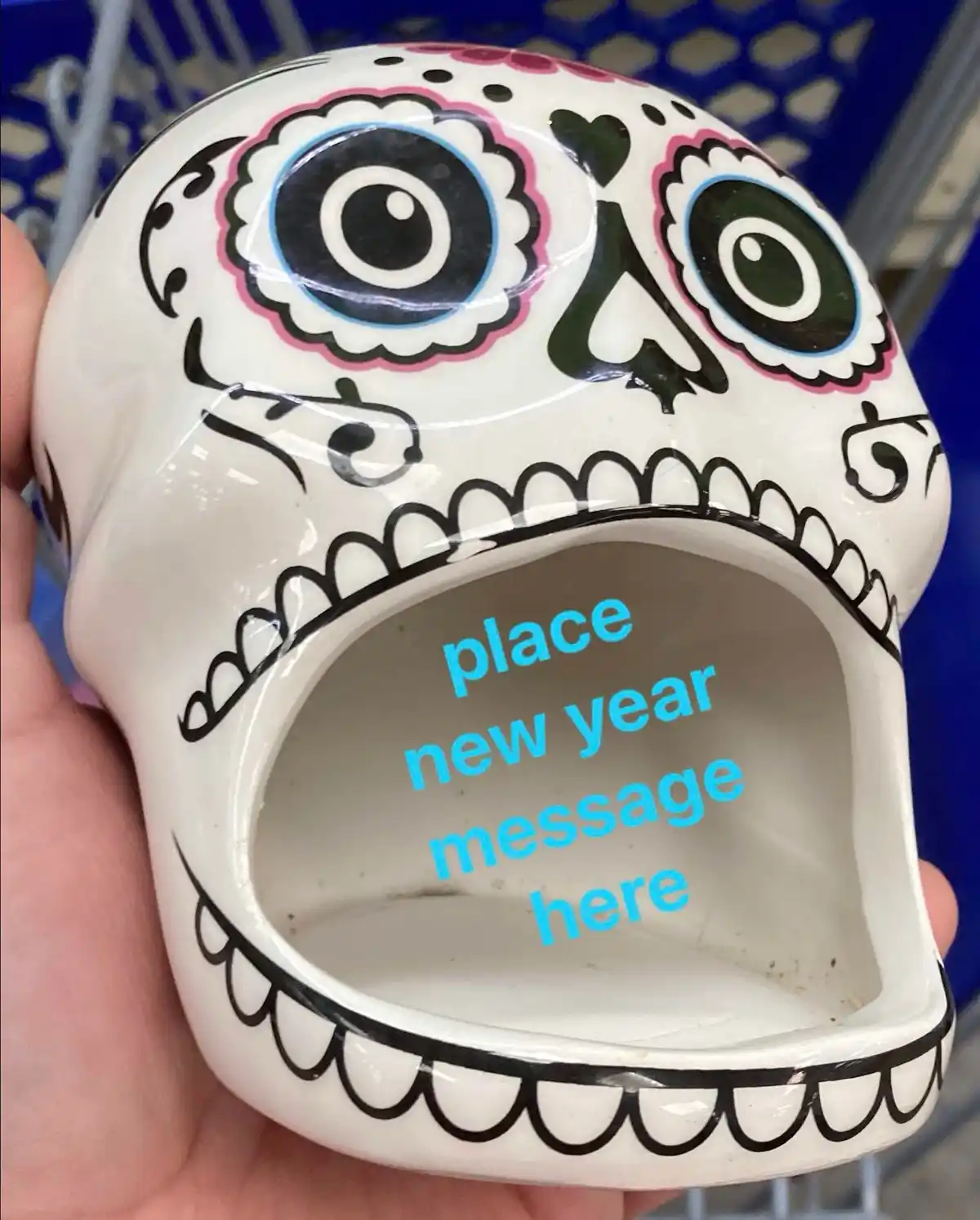A hand holds a white ceramic sugar skull decorated with colorful floral patterns around the eyes and black outlines, showing…