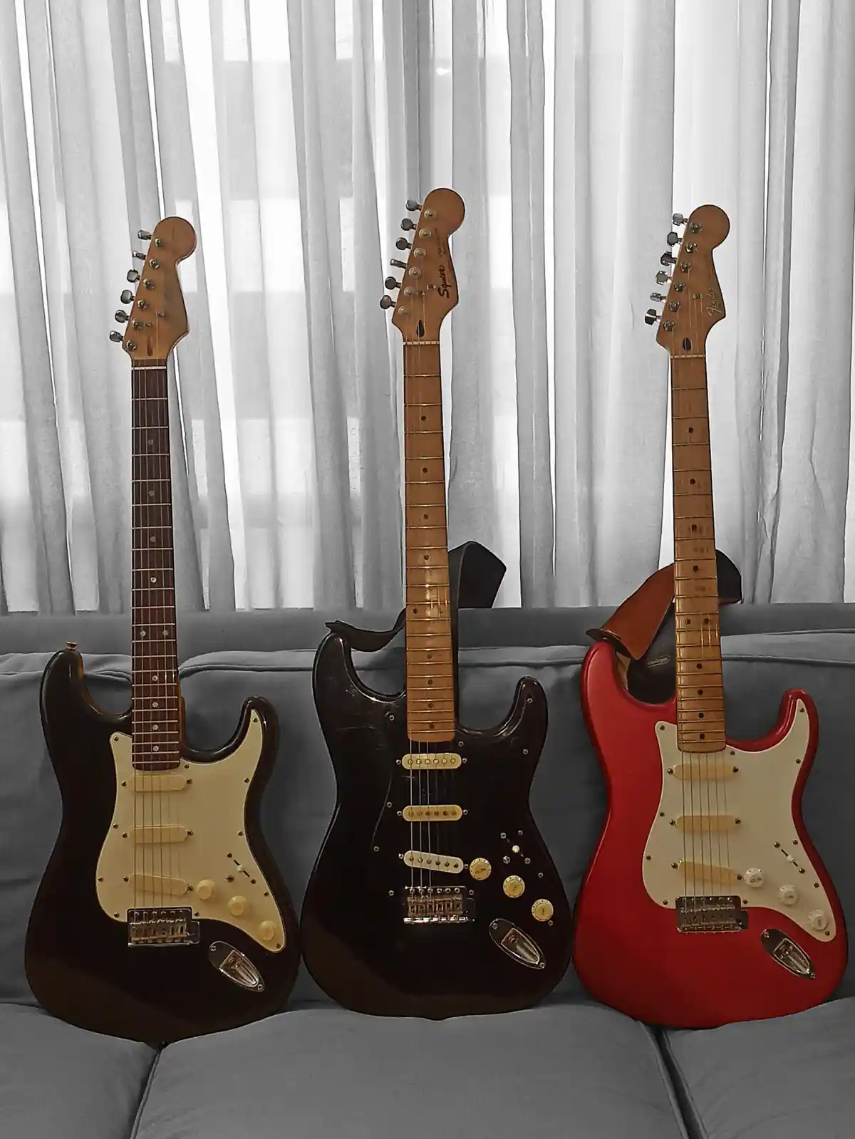 Three electric guitars, two dark and one red, are standing upright on a gray couch against a white curtain backdrop. The…