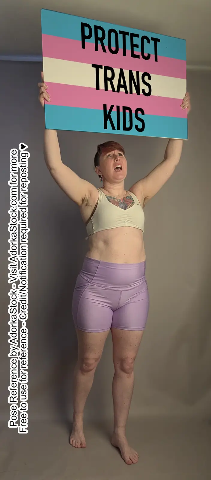 A person with short hair and visible tattoos holds up a sign with the trans flag colors and the text 'PROTECT TRANS KIDS'…