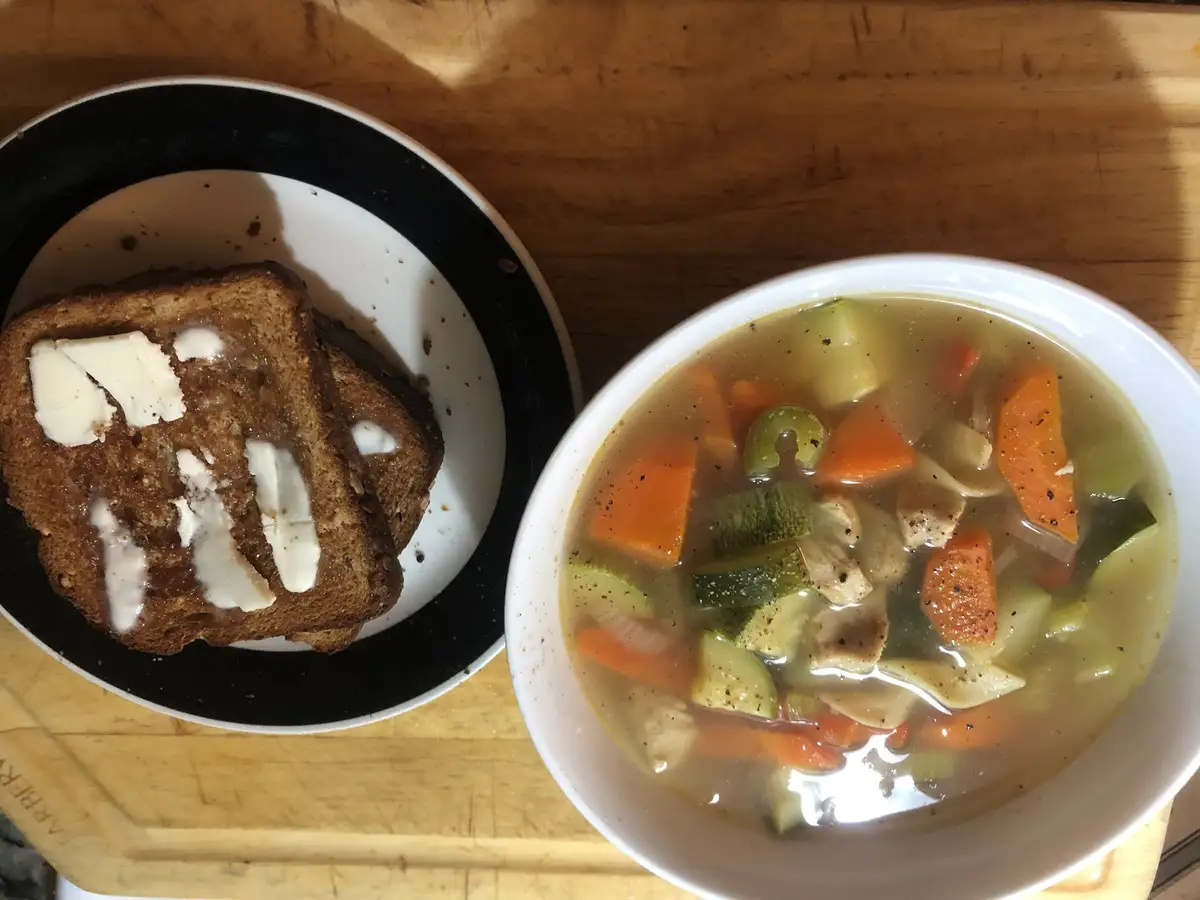 A top-down photo shows a white bowl of vegetable soup with carrots, zucchini, and chicken, next to a black-rimmed plate with…