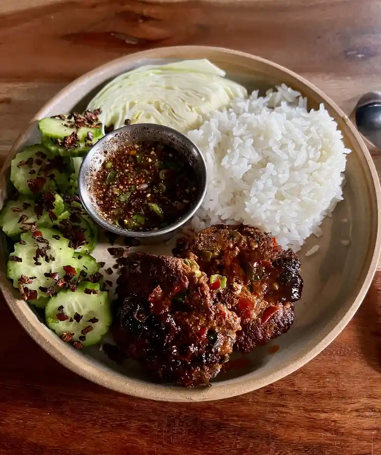 A close-up photo of a plate of food on a wooden table, featuring white rice, sliced cabbage, sliced cucumbers topped with…
