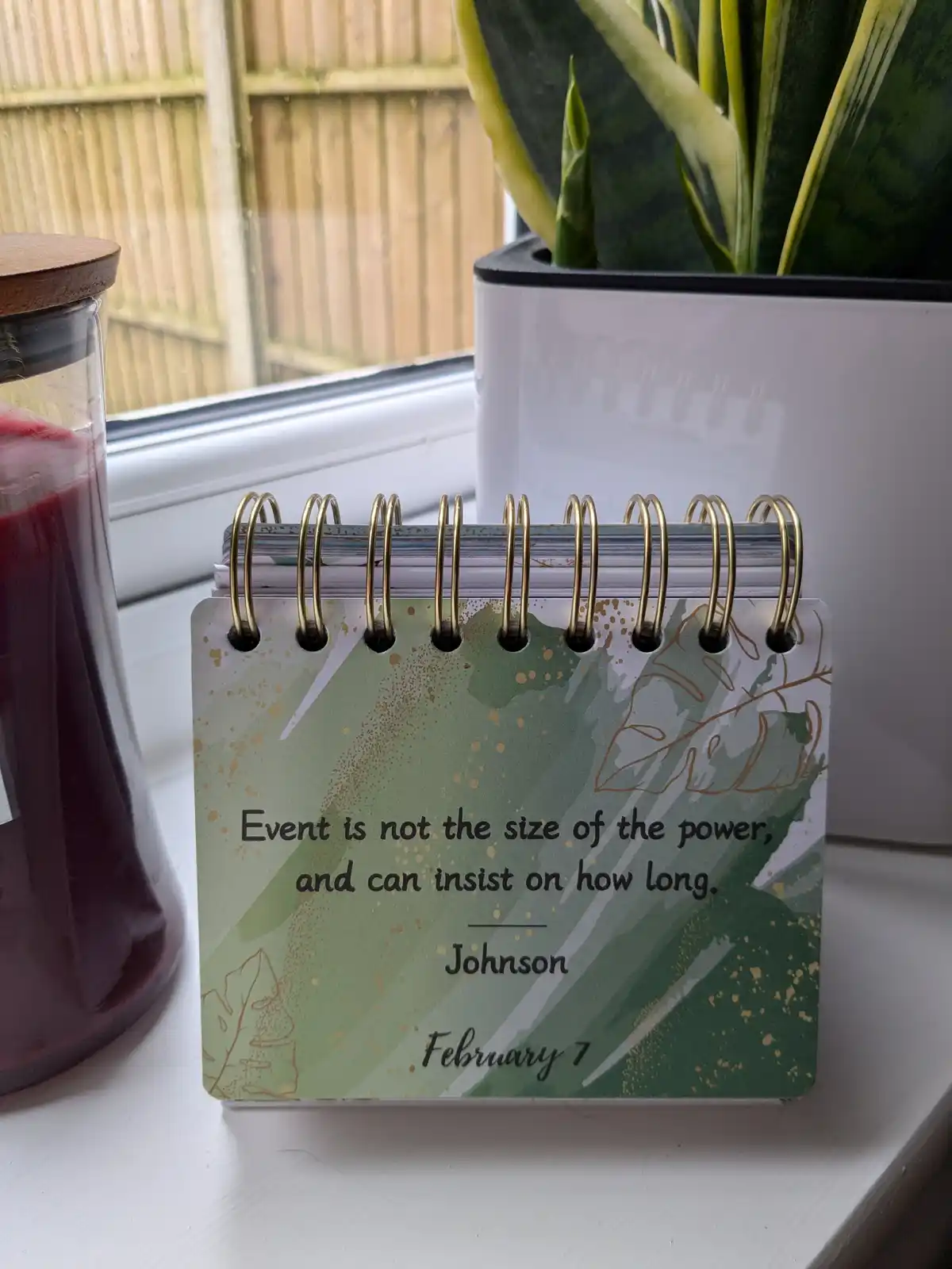 A spiral-bound desk calendar with a green and gold leaf-patterned cover sits on a windowsill next to a potted snake plant…