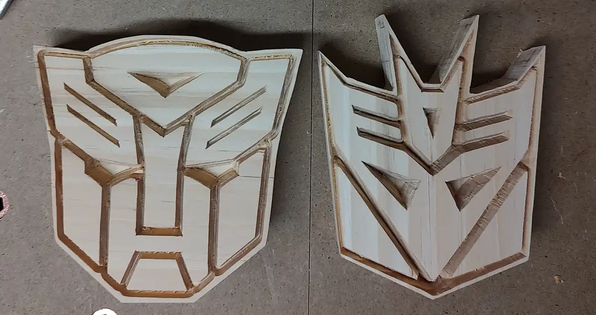 Two wooden carvings of the Transformers logos sit side by side on a work surface; the left is the Autobot symbol and the…