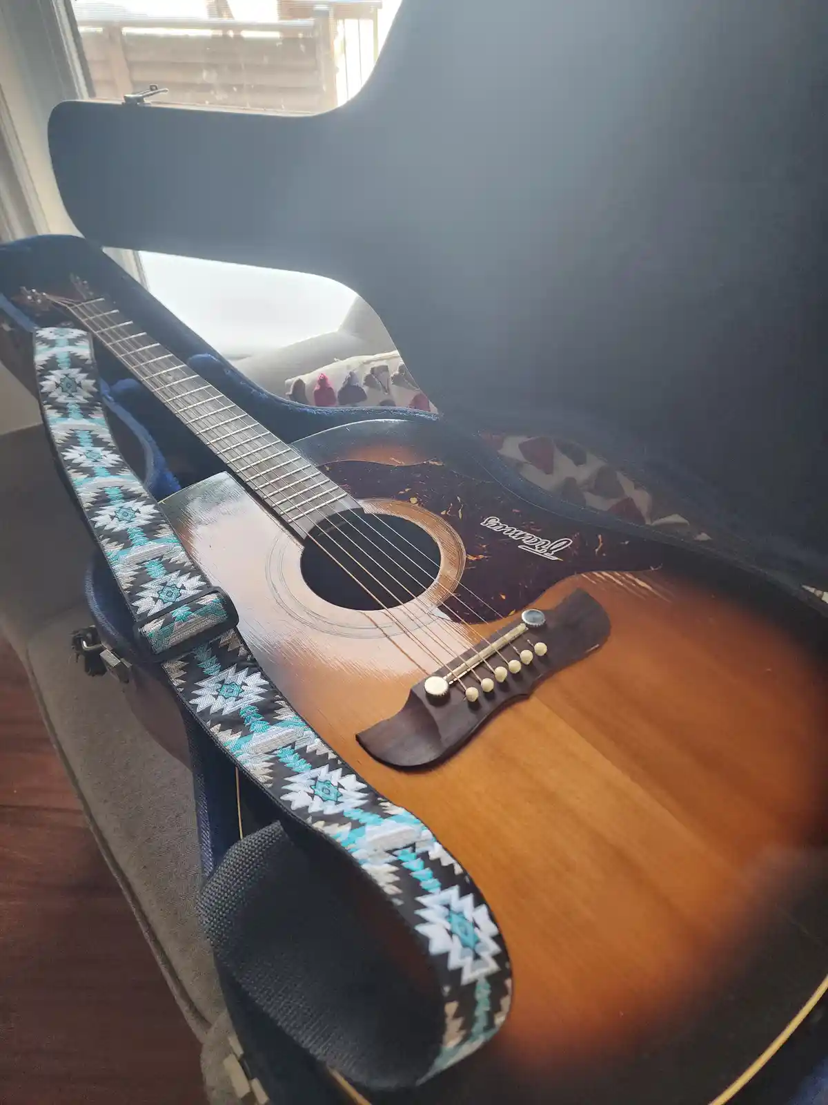 An acoustic guitar with a patterned strap rests inside its open black case, positioned near a window with natural light. The…