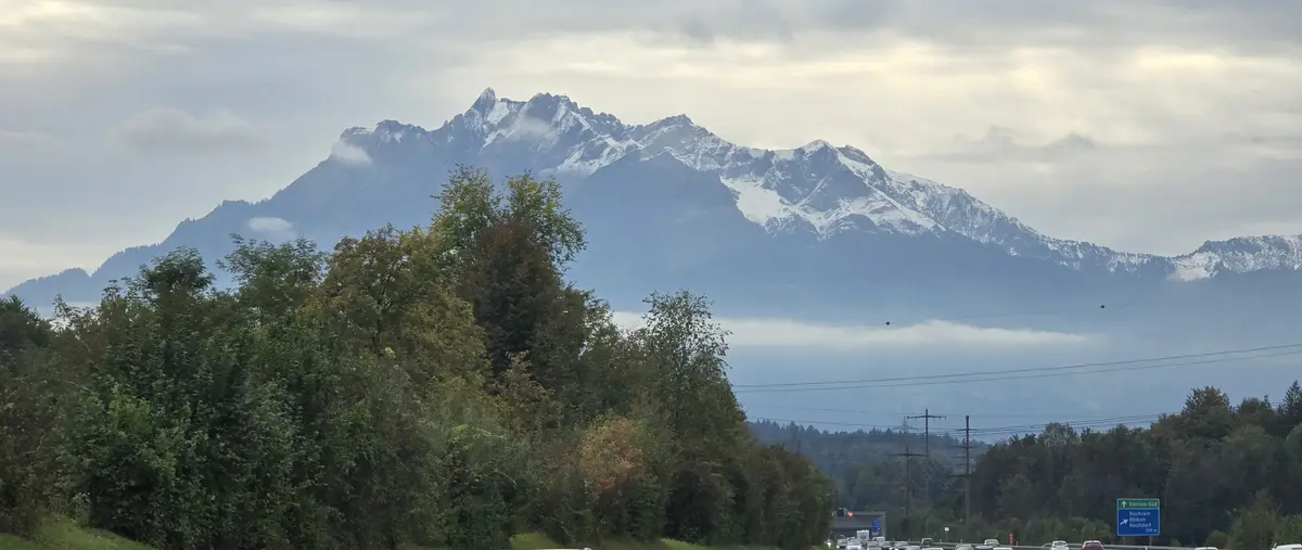A scenic view from a highway shows a majestic snow-capped mountain range under a cloudy sky, with green trees in the…