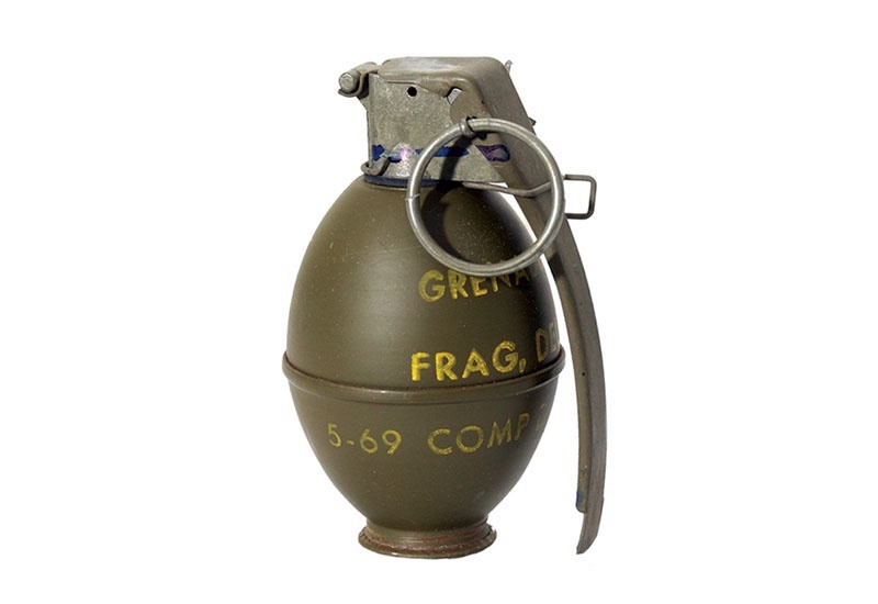 A military fragmentation hand grenade with a pull-ring and safety lever, showing faded yellow stenciled text reading…