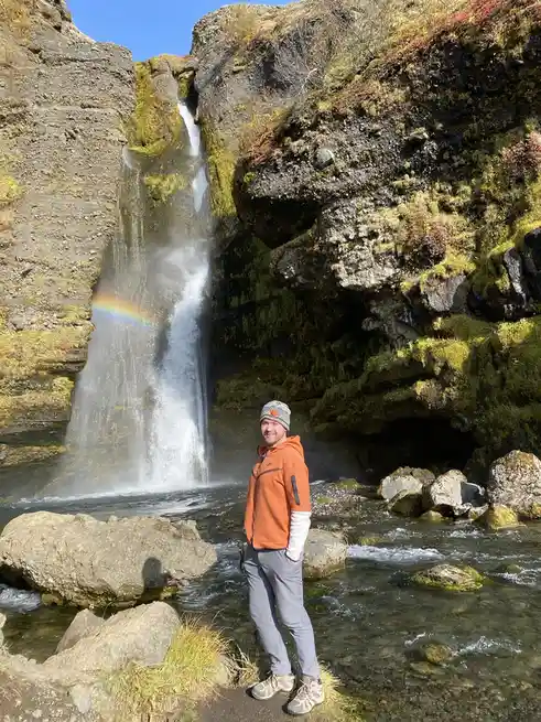 A man in an orange jacket and beanie stands smiling in front of a waterfall with a rainbow visible in the mist. The…