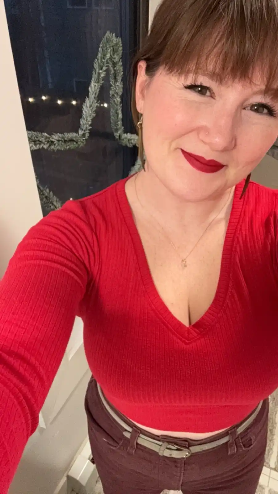 A woman with brown hair and red lipstick smiles at the camera while wearing a red ribbed crop top and dark pants. Behind…