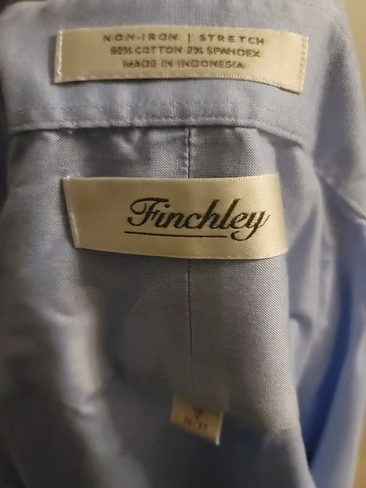 A close-up photo of the inside collar of a light gray shirt, showing three beige fabric tags. The top tag reads 'NON-IRON |…