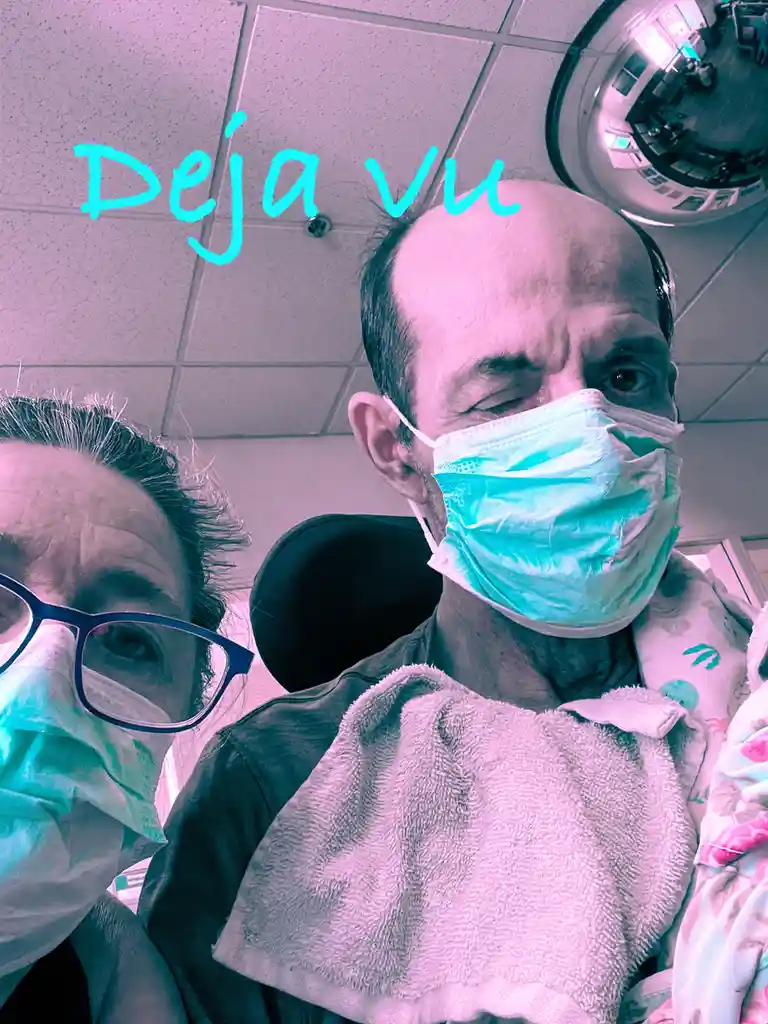 A selfie taken in a medical setting shows two people wearing surgical masks, one with glasses, both looking at the camera…