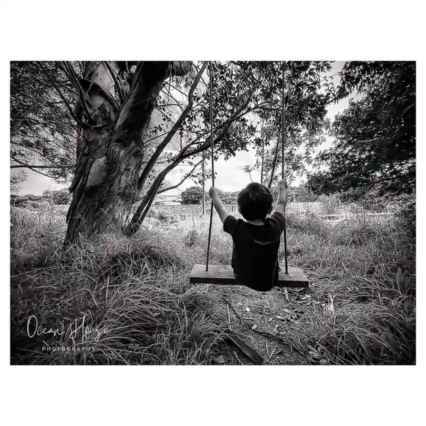 A black-and-white photo shows a young child sitting on a wooden swing, viewed from behind, with arms raised holding the…