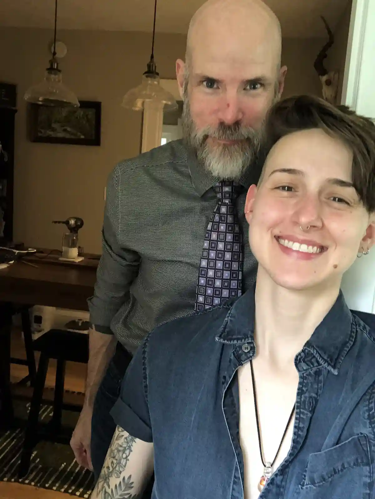 A selfie-style photo shows a smiling person with short hair and a denim shirt in the foreground, while a bald man with a…