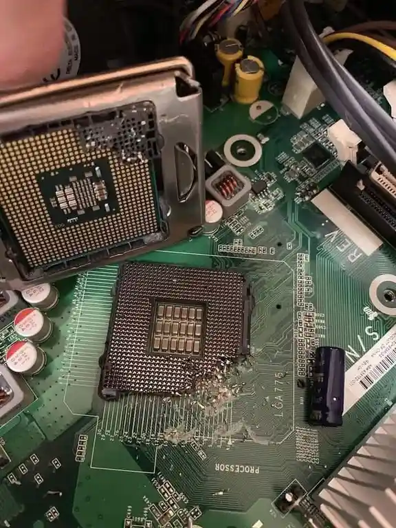 A close-up photo shows a damaged computer motherboard with a CPU socket that has shattered glass-like fragments around it. A…