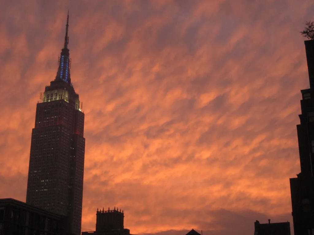 The Empire State Building stands tall against a dramatic, fiery orange sky filled with textured clouds, its upper floors and…