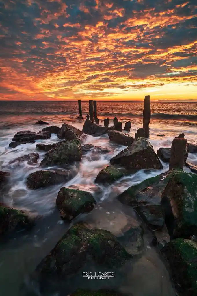 A long-exposure photograph captures waves flowing smoothly around moss-covered rocks at sunset, with vibrant orange and red…