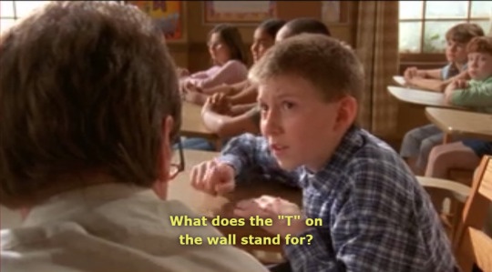 A young boy in a plaid shirt sits at a desk in a classroom, looking intently at his teacher who is facing away from the…