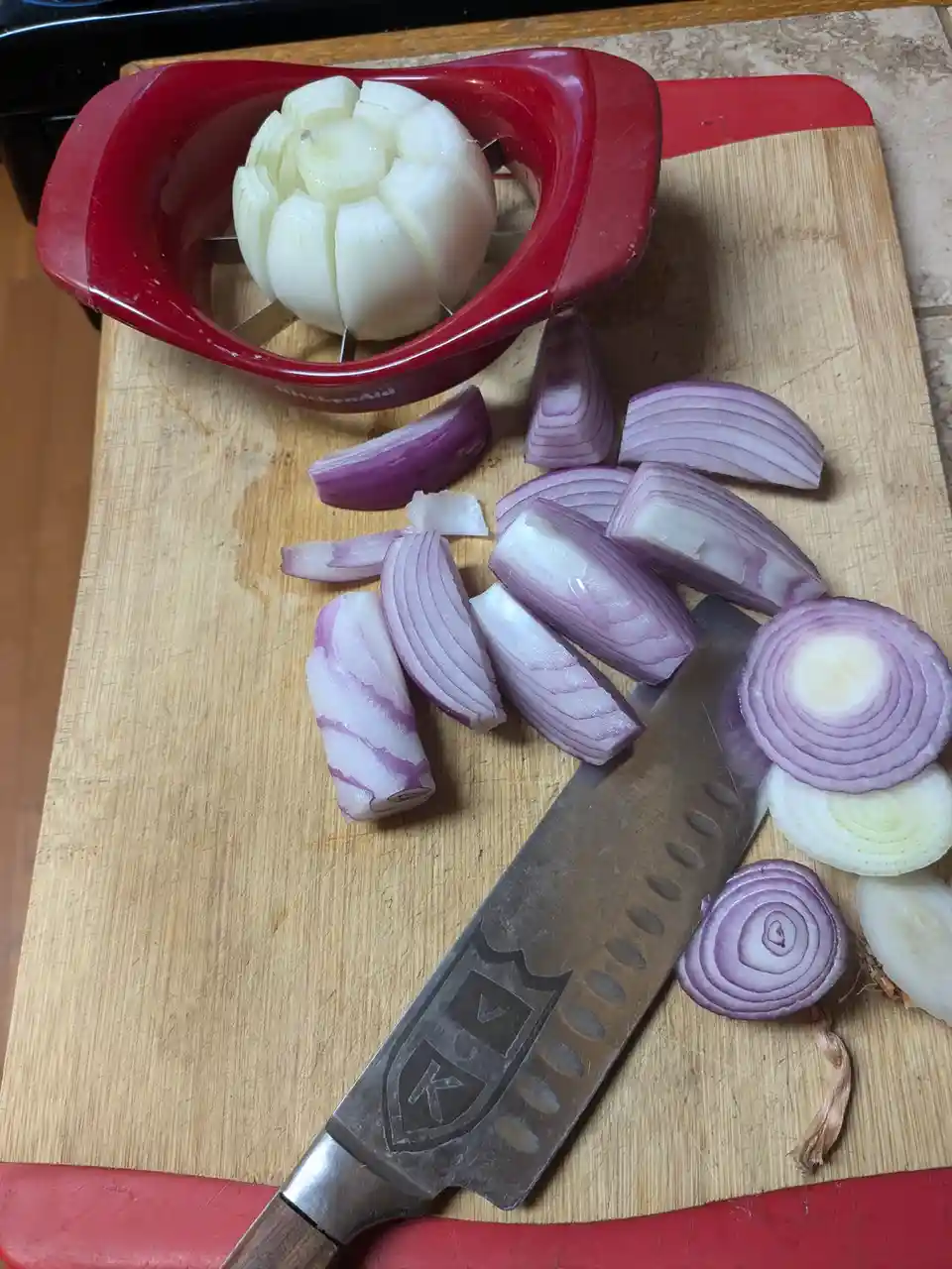 A wooden cutting board holds a red KitchenAid onion slicer with a white onion inside, surrounded by chopped red onions, a…
