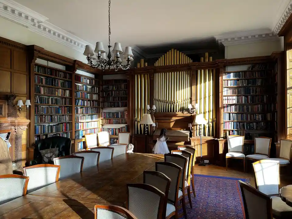 A young girl in a white dress stands in front of a large pipe organ in a grand, sunlit library filled with bookshelves…